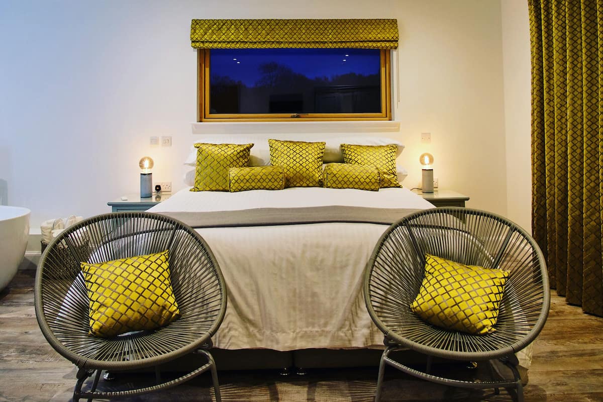 A bedroom with a bed, two chairs, a nightstand, and a window with a view of the sky.