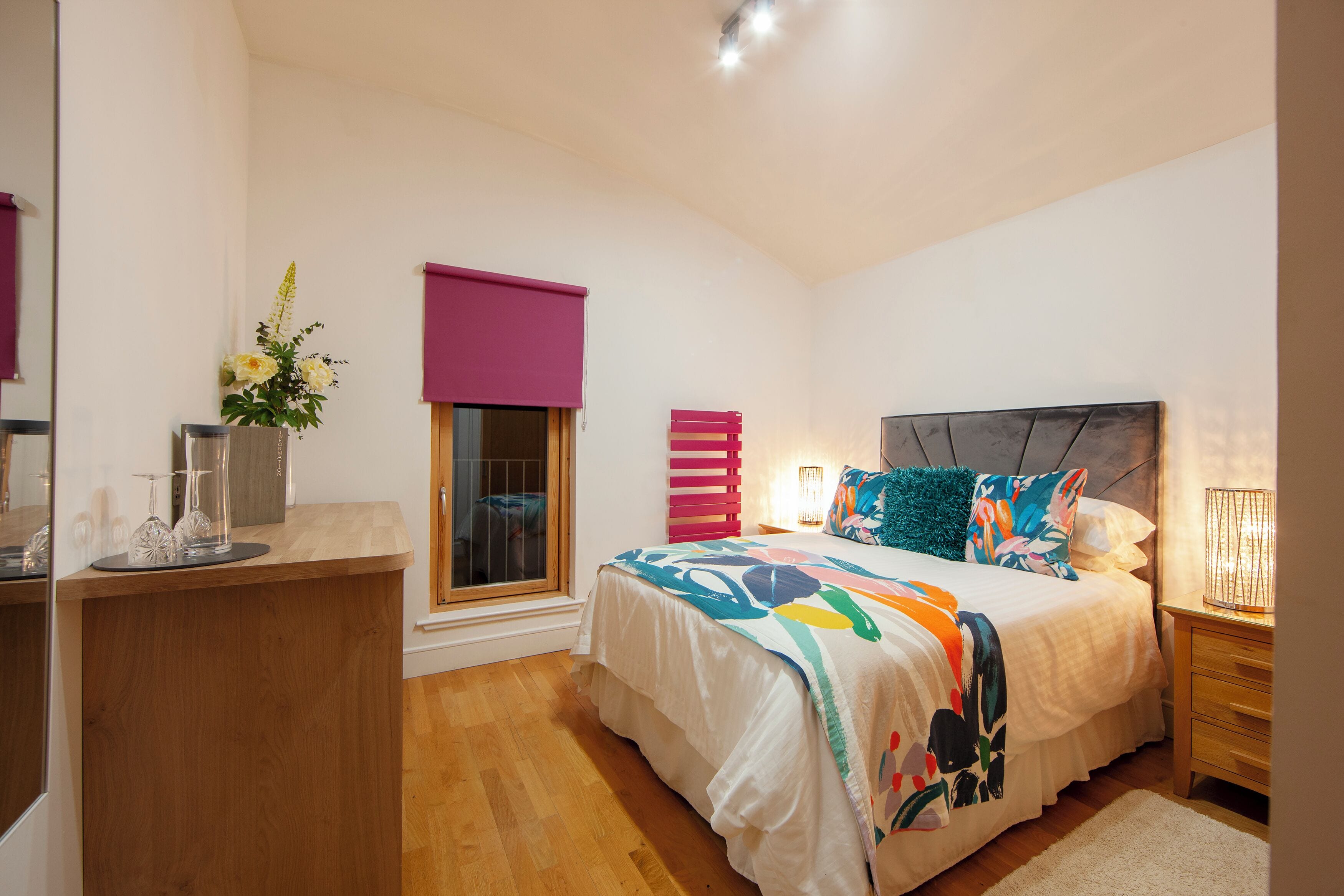 Une chambre à coucher comprenant un lit, une table de chevet, une fenêtre et un petit bureau agrémenté d’un vase de fleurs.