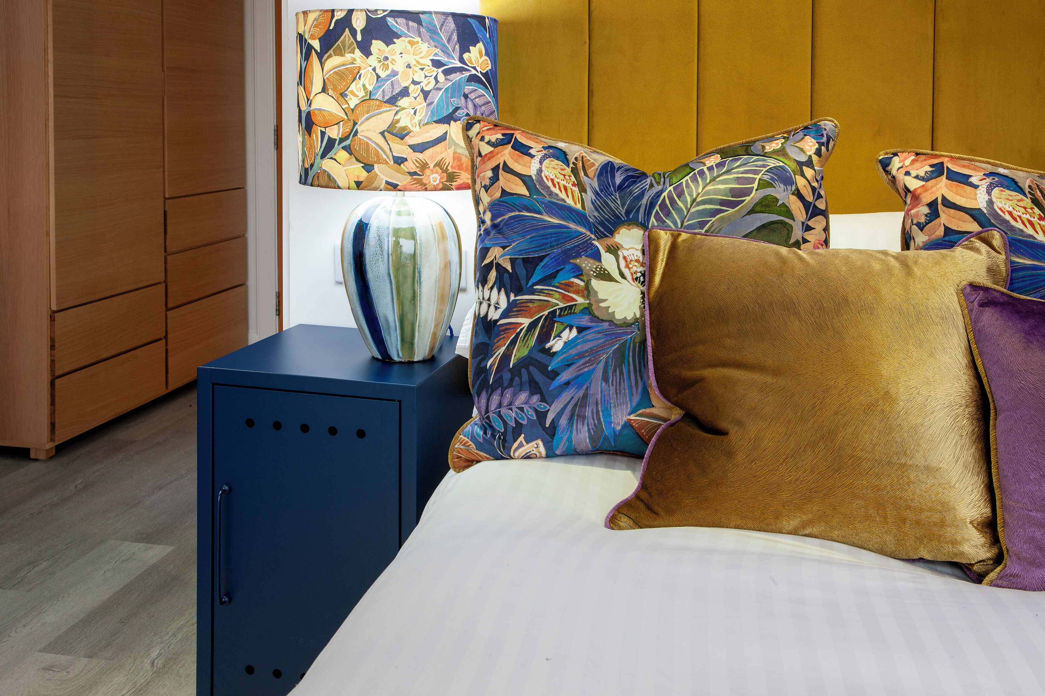 Una cama con almohadas estampadas con motivos florales, una mesita de noche con un florero y un armario de madera.