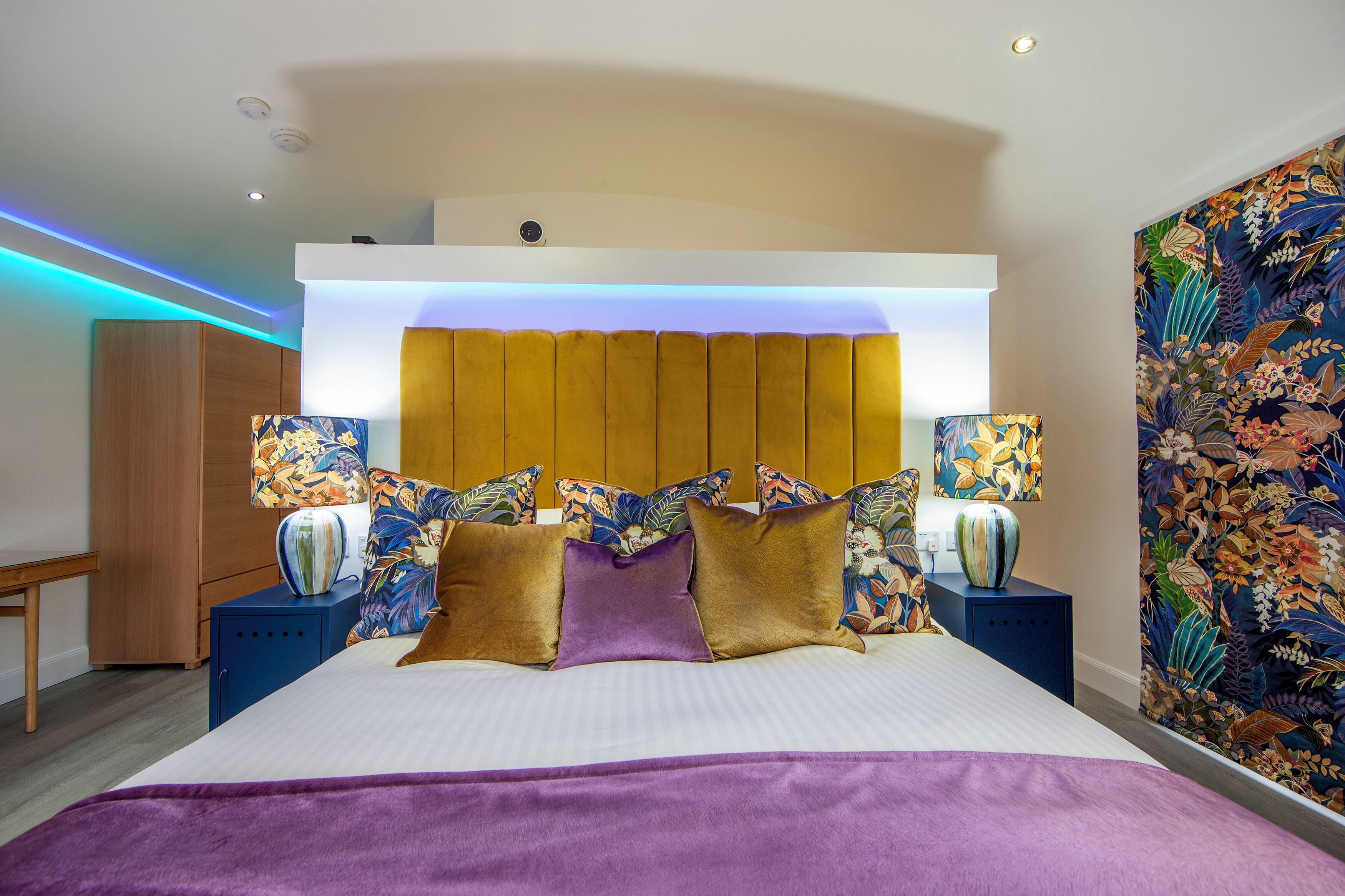 A hotel room with a large bed, a wooden headboard, two bedside tables with lamps, and a floral-patterned wall.