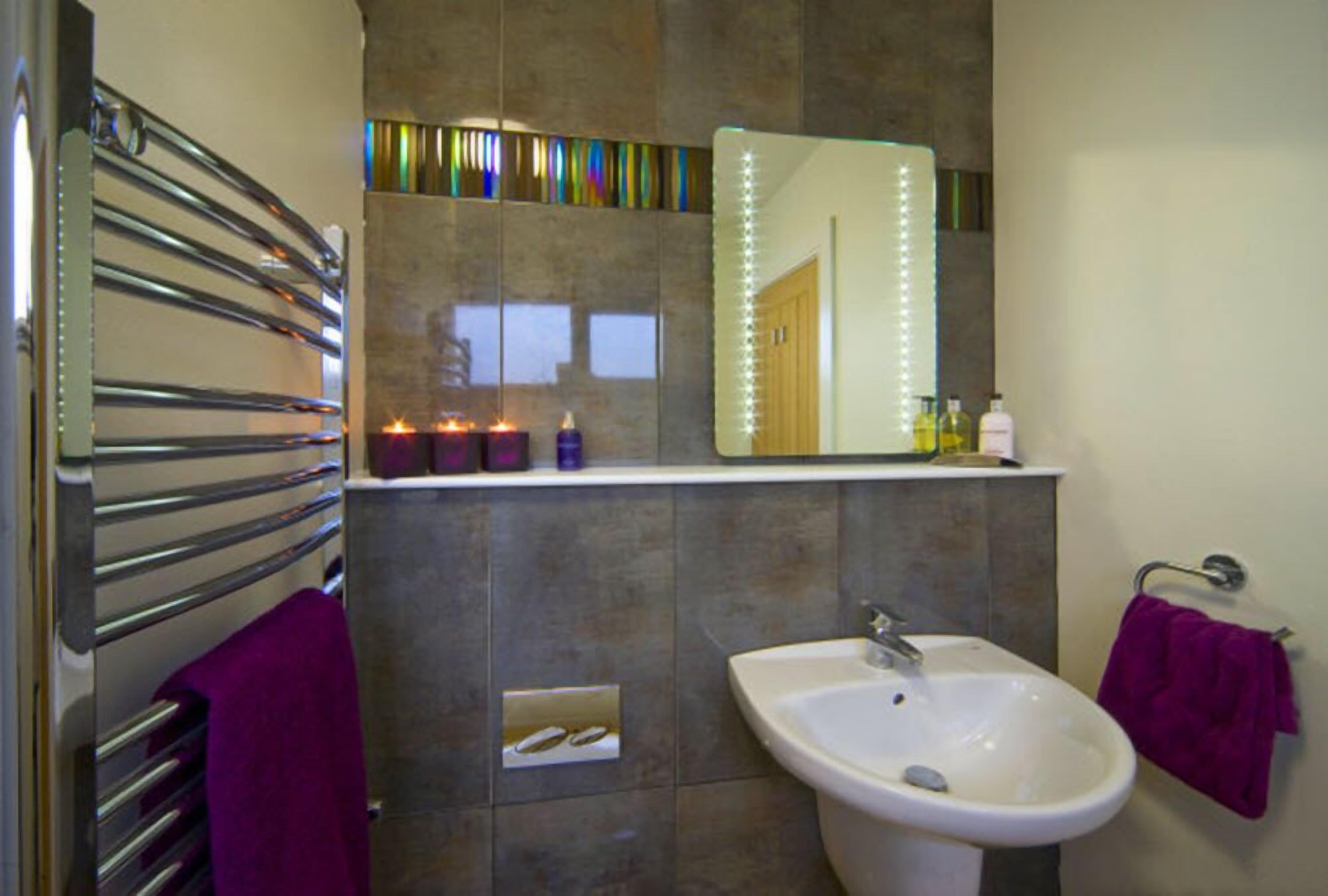 A bathroom with a white sink, a mirror with lights, a towel rack, and a shelf with toiletries.