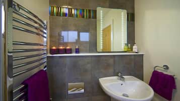 A bathroom with a white sink, a mirror with lights, a towel rack, and a shelf with toiletries.