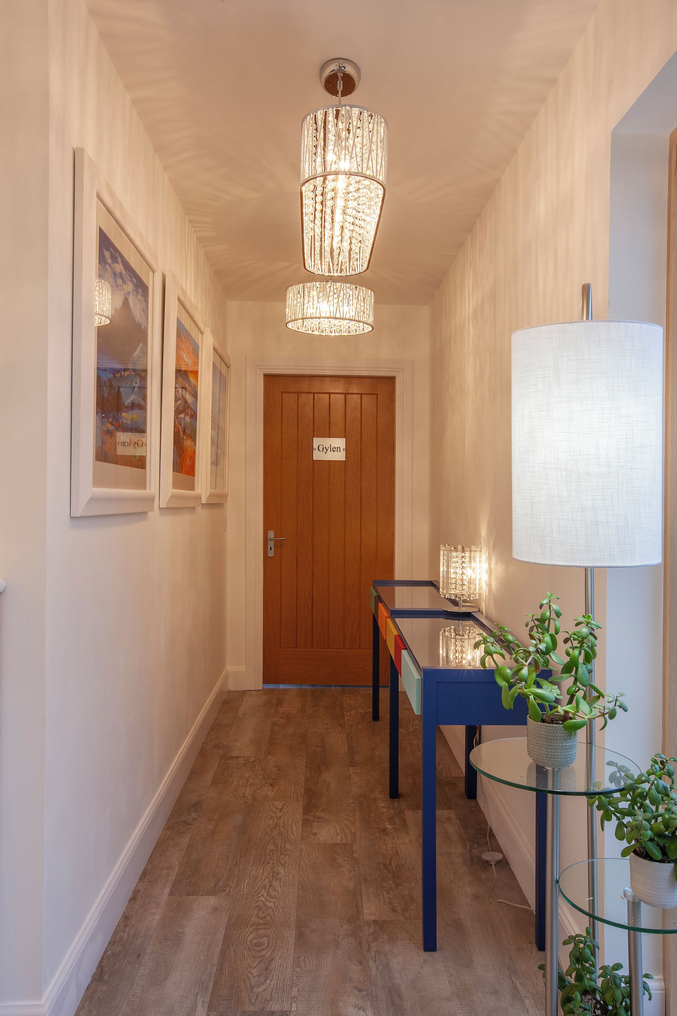 A hallway with a wooden door labeled "Gylle," a glass table with a blue base, and a floor lamp with a white shade.