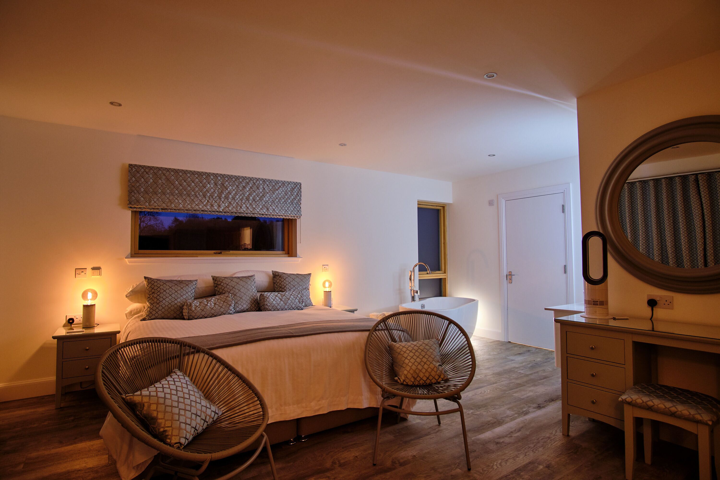 A bedroom with a large bed, two bedside tables, a nightstand, a chair, a dresser, a mirror, and a window with a view.