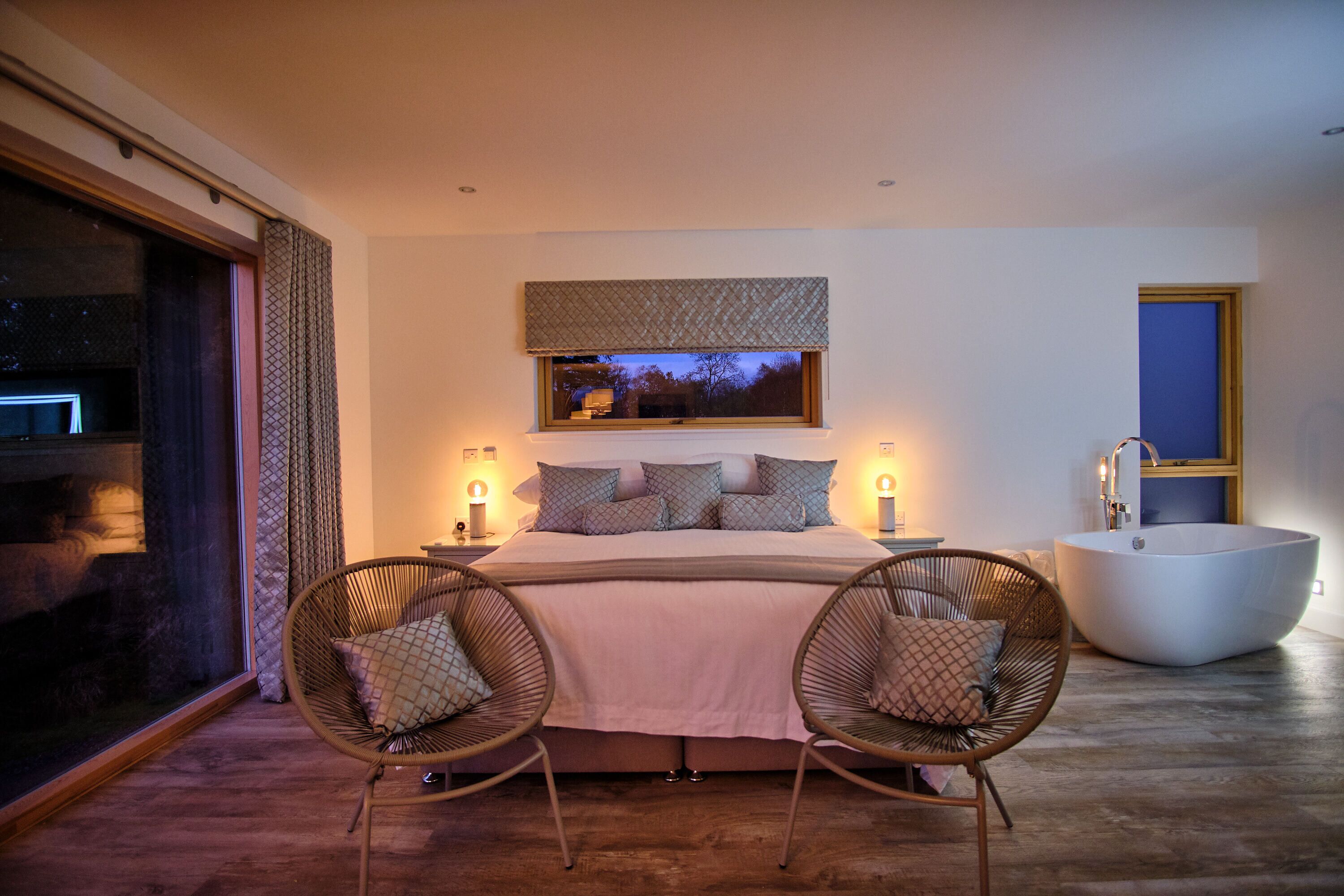 A bedroom with a large bed, two wicker chairs, a bathtub, and a window with a view.