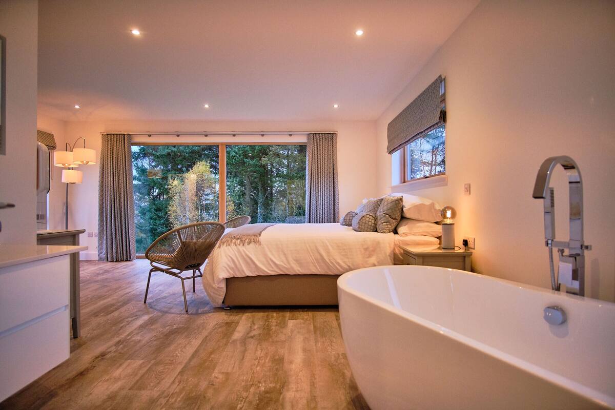 A modern bedroom with a large bed, a sitting area with a chair, a bathtub, and a view of trees through the window.