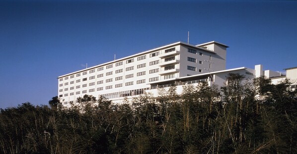Un edificio de varios pisos con techo plano y ventanas rectangulares, rodeado de árboles frondosos.
