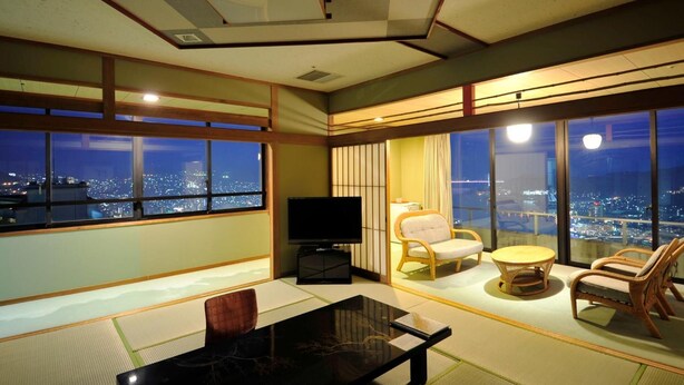 Une chambre d’hôtel moderne avec un sol recouvert de tatamis, un téléviseur à écran plat, une table à manger et une vue sur la ville la nuit.