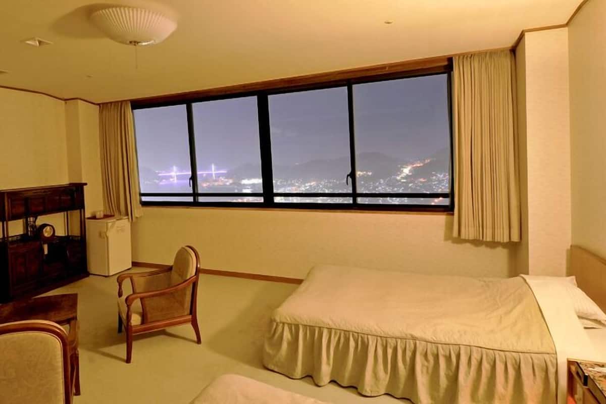 Ein Hotelzimmer mit großem Fenster, einem Bett, einem Sessel, einem Schreibtisch und einer Kommode.