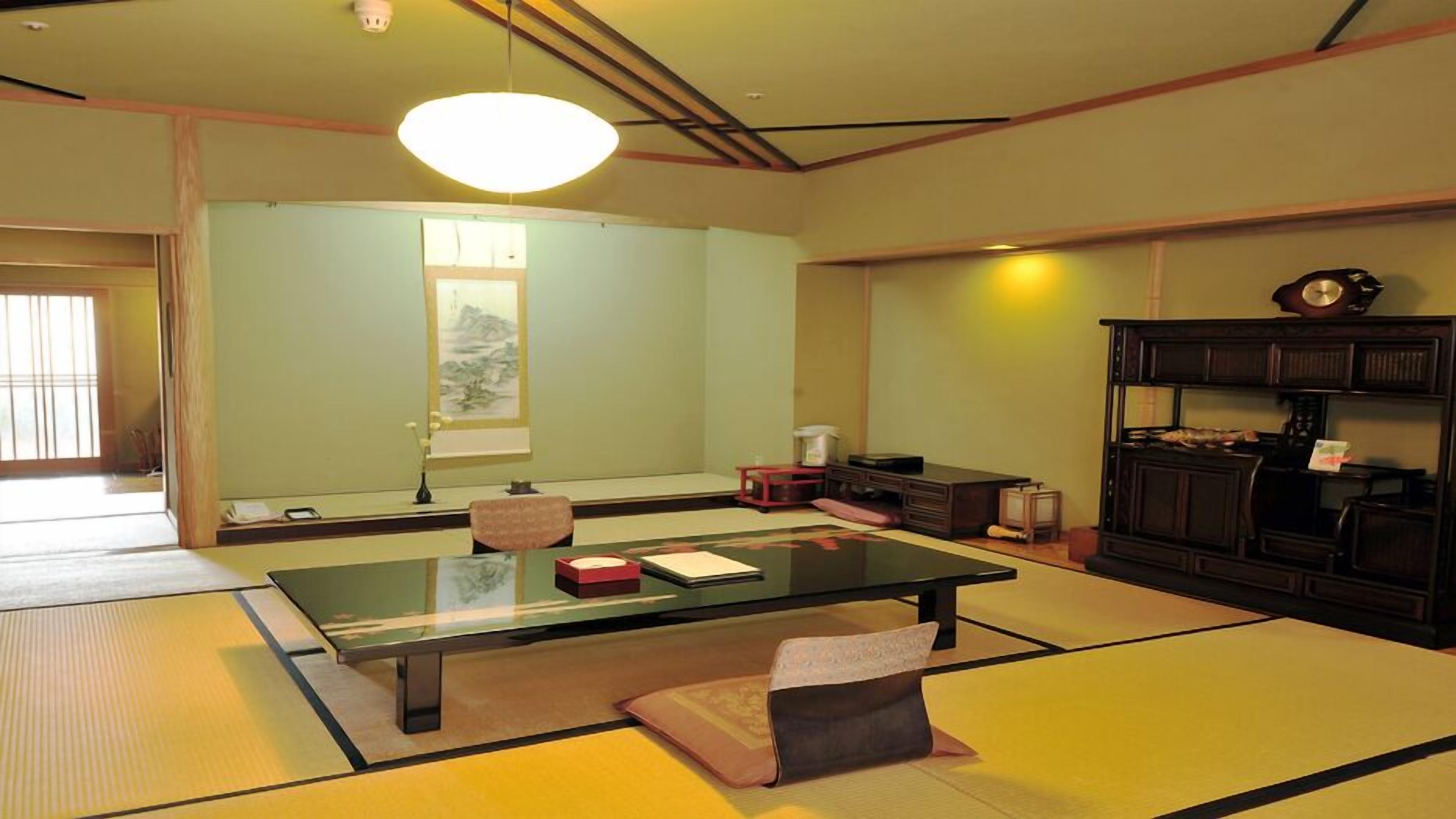 Une pièce avec un sol recouvert de tatamis, une petite table basse, des coussins, une peinture traditionnelle japonaise et un buffet en bois foncé.