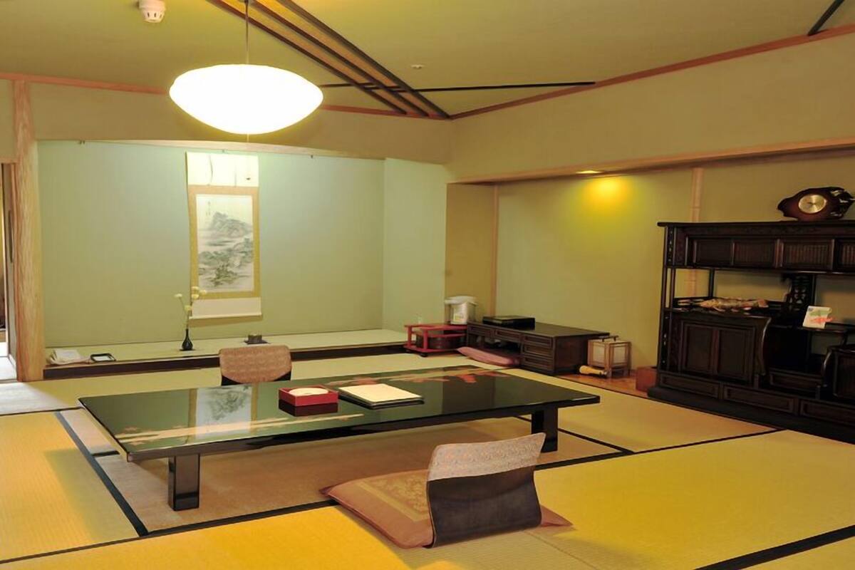 Ein Raum mit Tatami-Matten, einem niedrigen Tisch, Kissen, einem traditionellen japanischen Gemälde und einer dunkelholzigen Kommode.