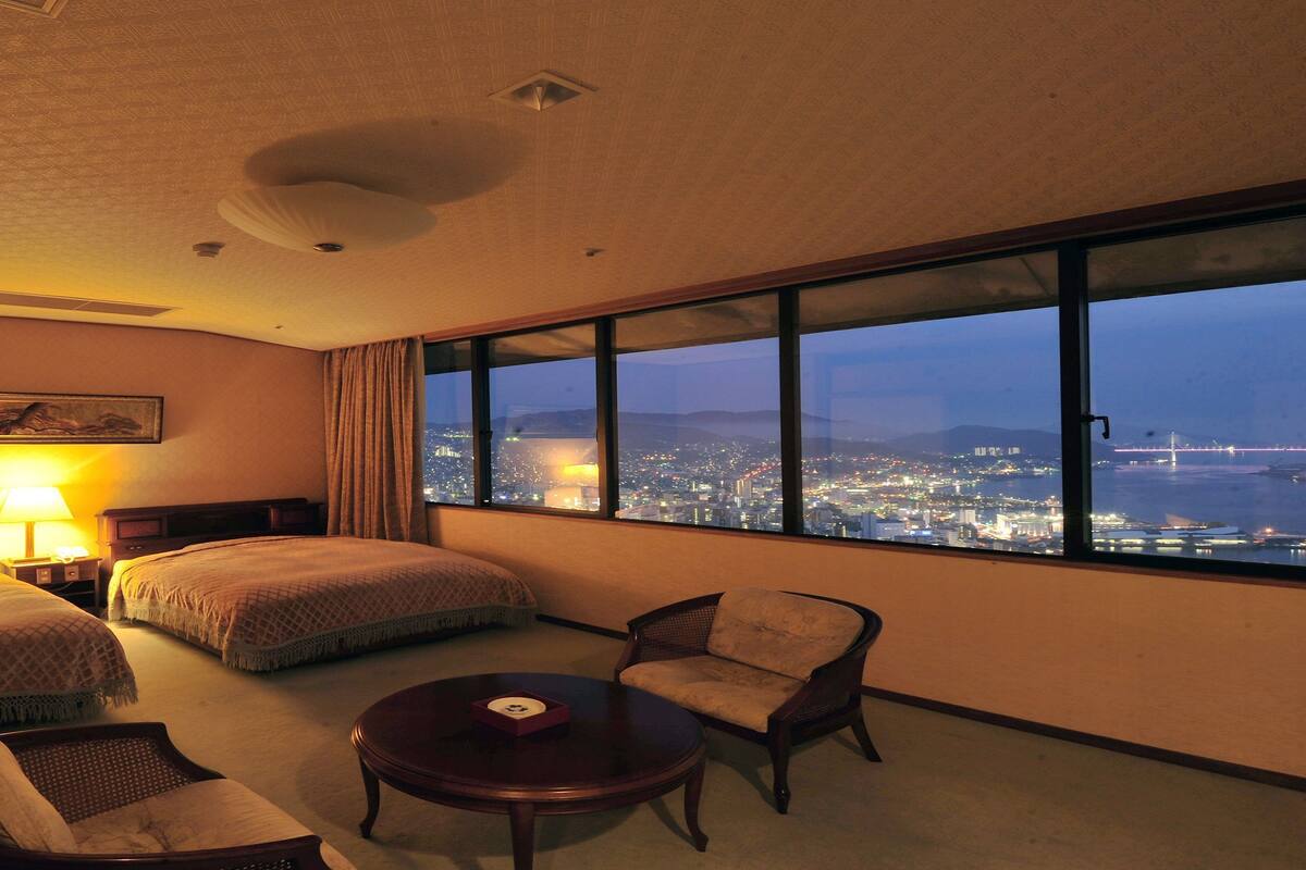 Ein Hotelzimmer mit zwei Betten, einem Sessel, einem Tisch und einem großen Fenster mit Blick auf die beleuchtete Stadt bei Nacht.