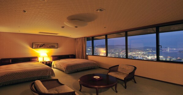Habitación de hotel con dos camas, una silla, una mesa y un amplio ventanal con vistas a la ciudad de noche.