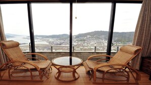 Un balcon avec des fauteuils en osier et une table en verre, offrant une vue sur un paysage urbain avec des montagnes et une étendue d’eau.