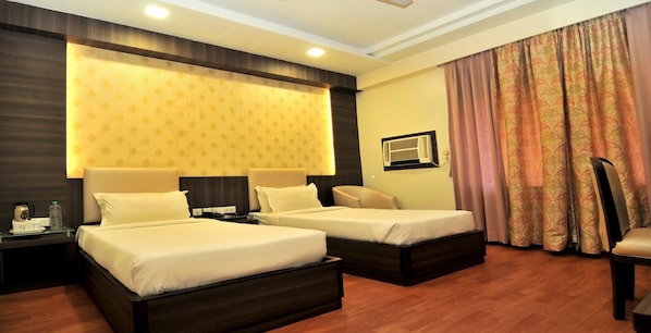 A hotel room with two beds, a bedside table, a chair, a large window with curtains, and a wall-mounted air conditioning unit.