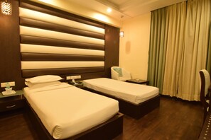 A hotel room with two single beds, a bedside table with a lamp, a chair, and a window with curtains. - Sojourn (Kolkata)