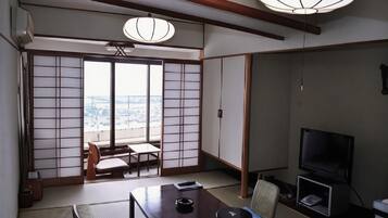 A traditional Japanese room with tatami flooring, a low table, a television, and a sliding door leading to a balcony with a city view.