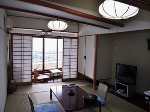 A traditional Japanese room with tatami flooring, a low table, a television, and a sliding door leading to a balcony with a city view. - Atami Tamanoyu Hotel (Shizuoka)