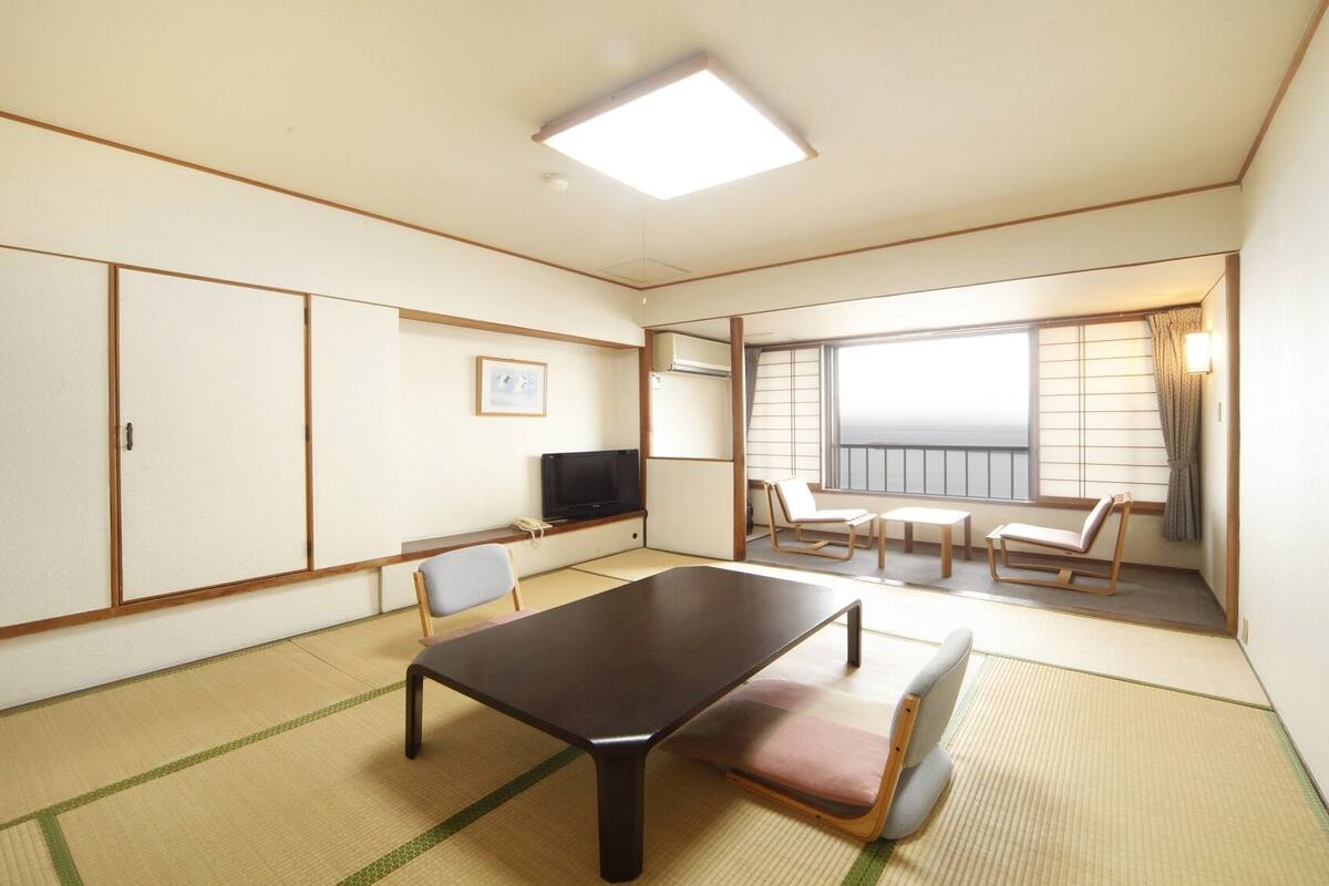 Ein traditionell japanisch gestalteter Raum mit Tatami-Boden, einem niedrigen Tisch, Stühlen, einem Fernseher und einem großen Fenster mit Blick.