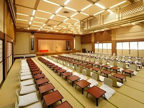 A conference room with rows of tables and chairs, a stage with a curtain, and a traditional Japanese aesthetic. - Atami Tamanoyu Hotel (Shizuoka)