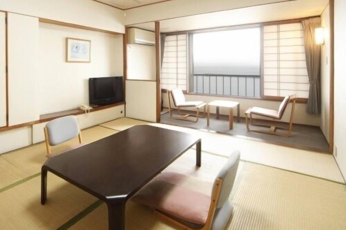 Ein Hotelzimmer mit Tatami-Böden, einem dunklen Holztisch, einem Fernseher, einer Couch und Meerblick durch große Fenster.