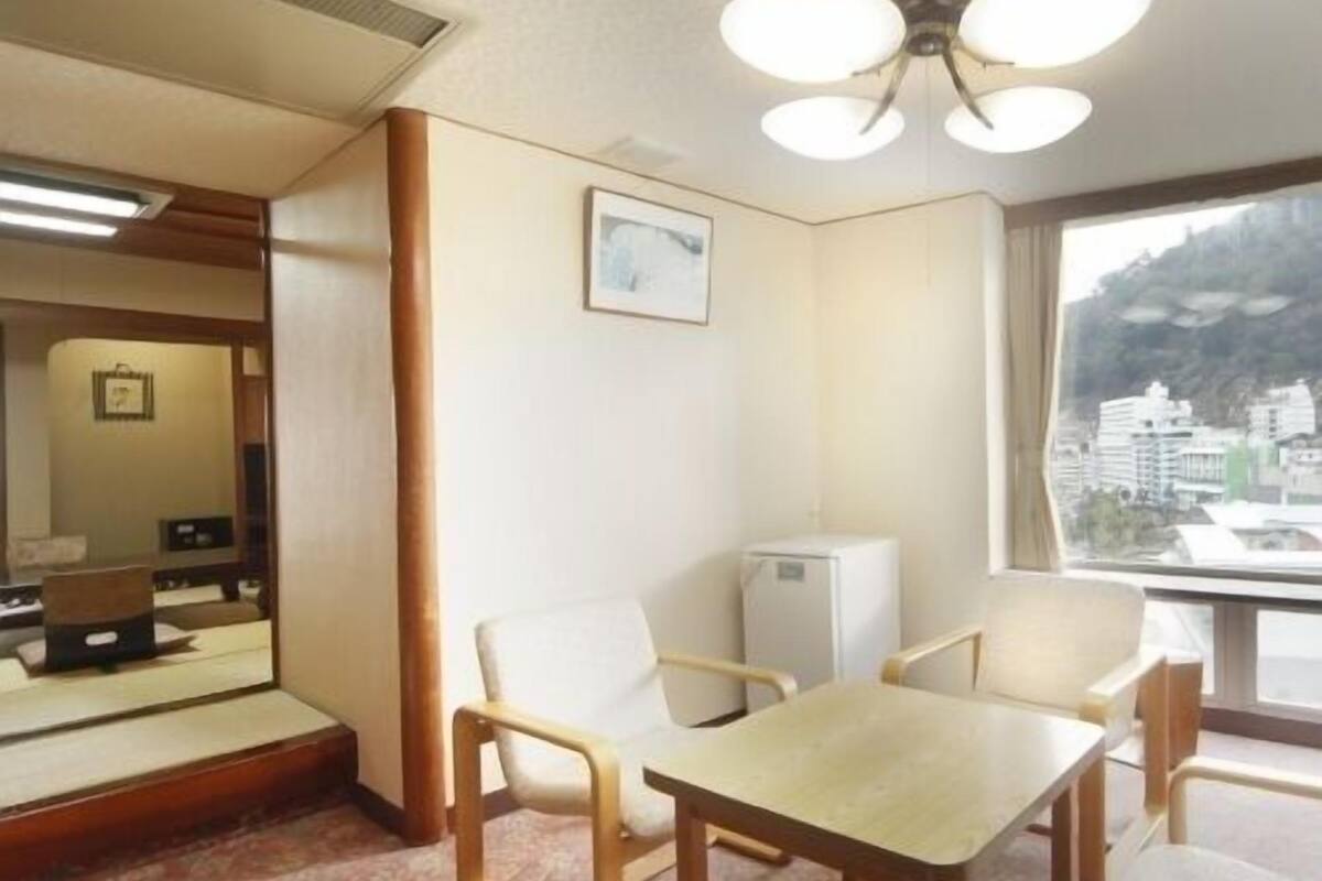 Ein Zimmer mit einem Holztisch, zwei Stühlen, einem kleinen weißen Kühlschrank, einem gerahmten Bild an der Wand und einem Fenster mit Blick auf Gebäude und Hügel.