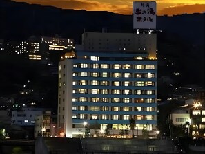 A multi-story hotel building with illuminated windows, a sign with Japanese characters, and a backdrop of a sunset over a mountainous area. - Atami Tamanoyu Hotel (Shizuoka)