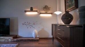 Un salon moderne avec un téléviseur, un plancher en bois, un vase blanc contenant des fleurs rouges, un meuble en bois foncé et des luminaires fixés au mur.
