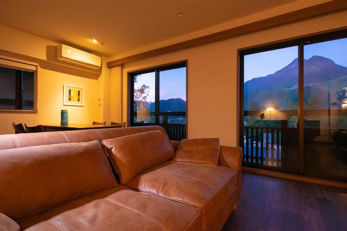 A spacious living area with a large leather sofa, a dining table, and a view of mountains through large sliding doors.