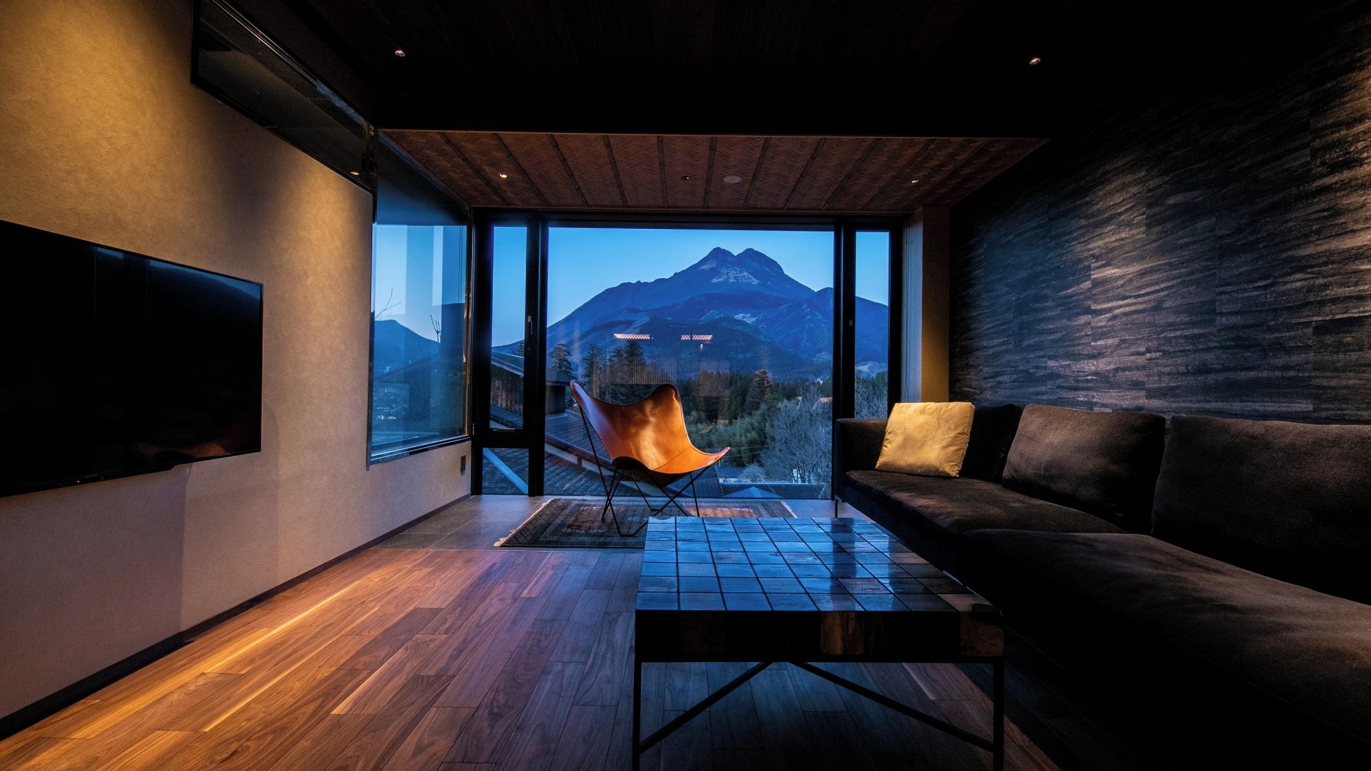 A modern living room with a large window, a flat-screen TV, a sofa, and a wooden floor.