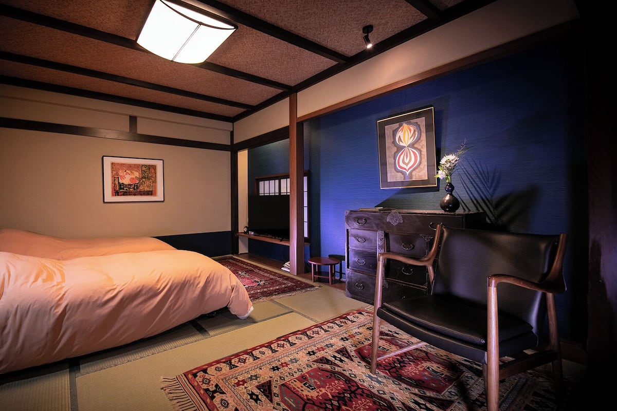 Une pièce de style japonais traditionnel, avec un sol recouvert de tatamis, un lit bas en bois, un bureau avec une chaise et un mur d’accent bleu.