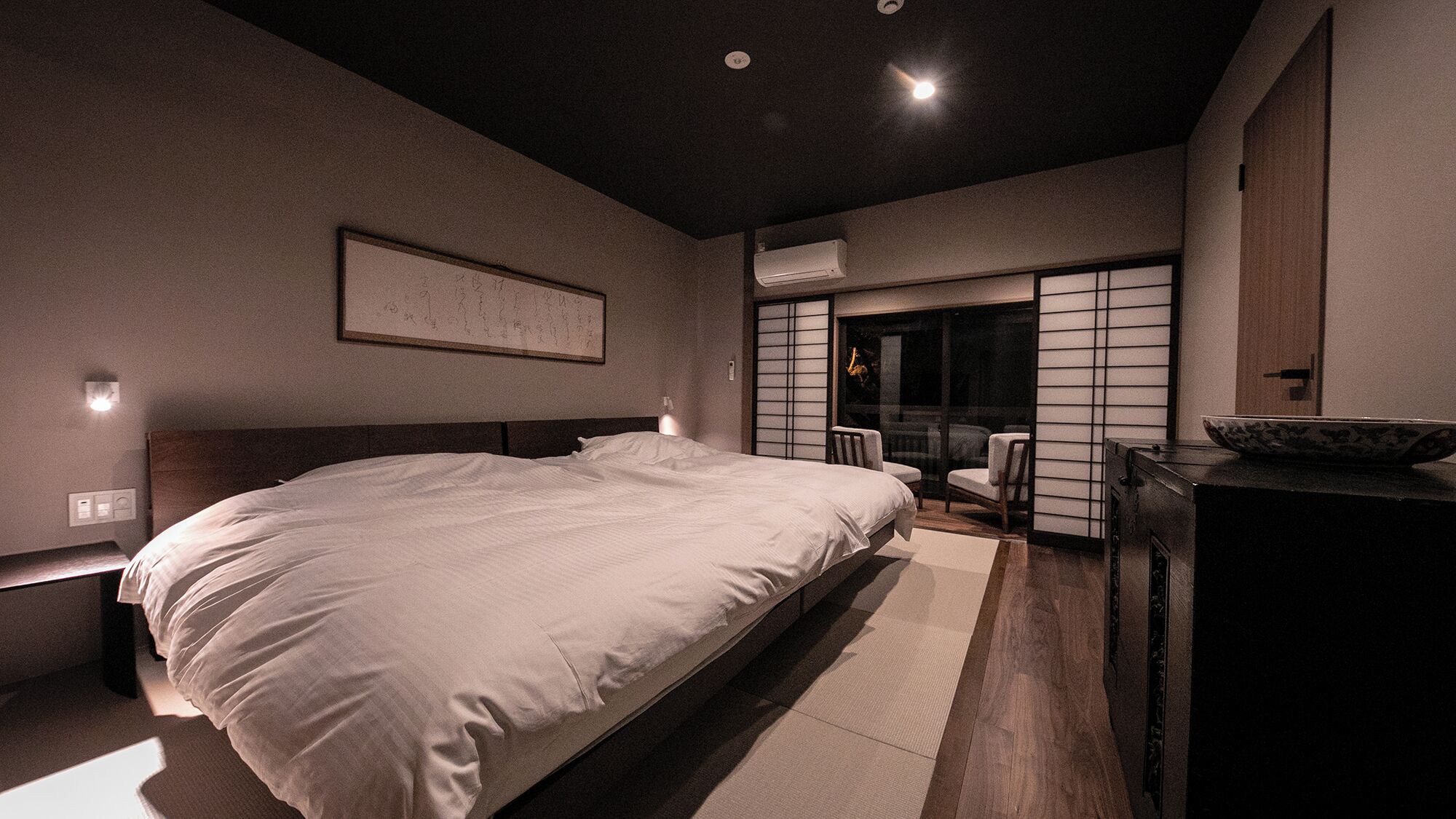 Een moderne hotelkamer met een groot bed, een bureau met een schaal, en een schuifdeur naar een zithoek.