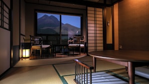 Een traditionele Japanse kamer met tatami vloerbedekking, een schuifdeur naar een balkon met uitzicht op bergen, en een tafel met stoelen.