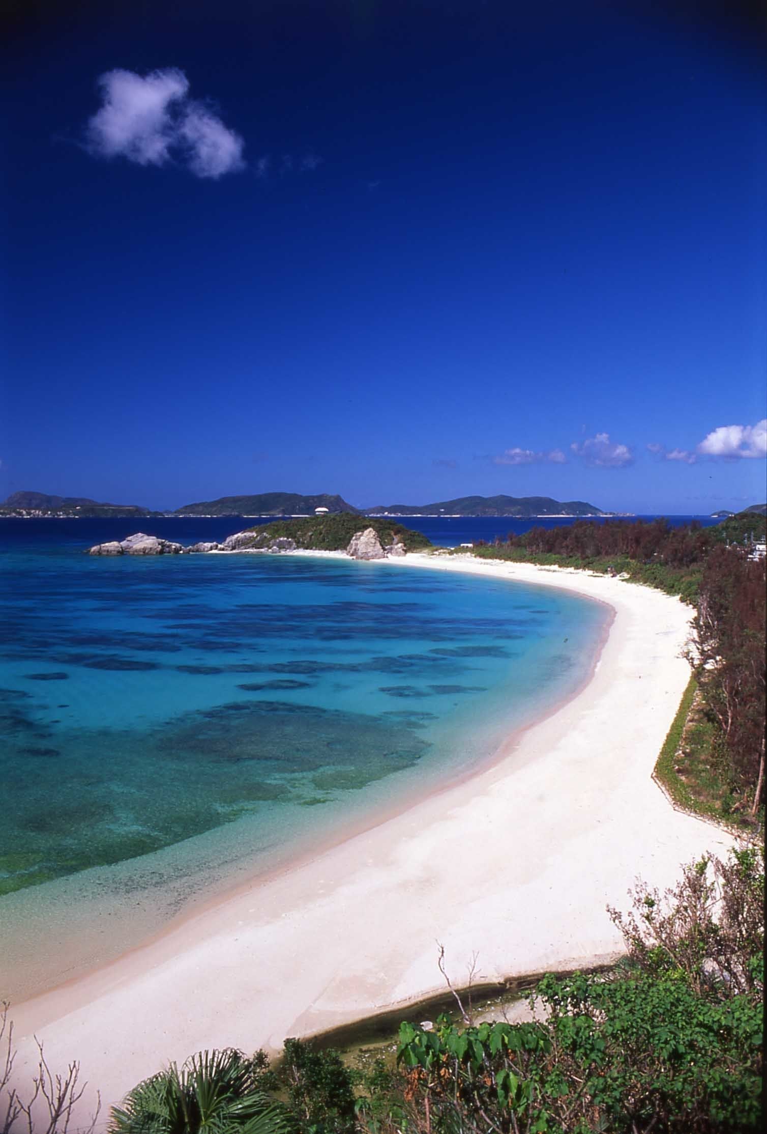 Una playa con aguas turquesas cristalinas, una orilla de arena blanca y colinas verdes al fondo.