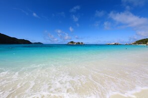 Una playa con agua turquesa cristalina, orilla arenosa e islas rocosas distantes bajo un cielo azul.