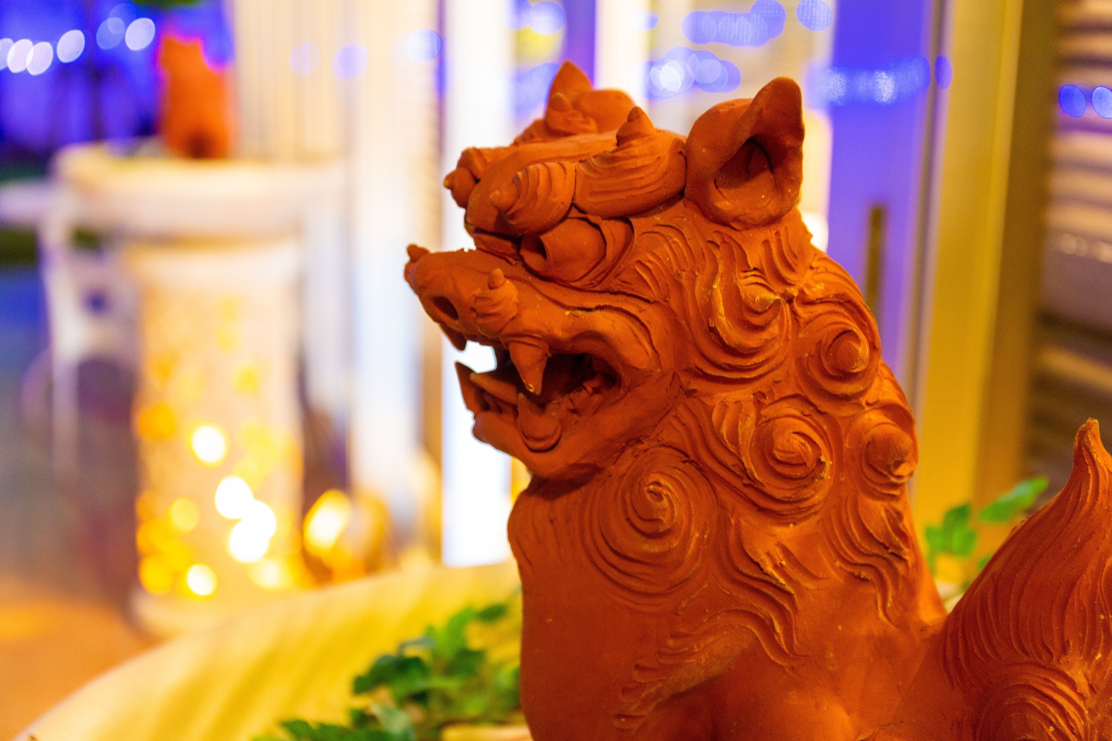 A terracotta lion statue with intricate carvings, placed on a table with a blurred background of indoor lighting and greenery.