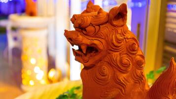 A terracotta lion statue with intricate carvings, placed on a table with a blurred background of indoor lighting and greenery.