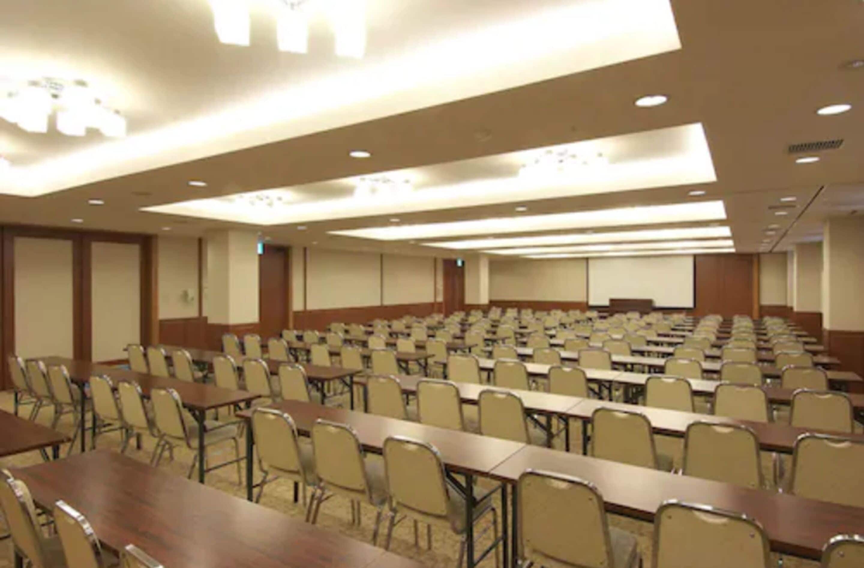 A conference room with rows of tables and chairs, a projector screen, and a stage area.