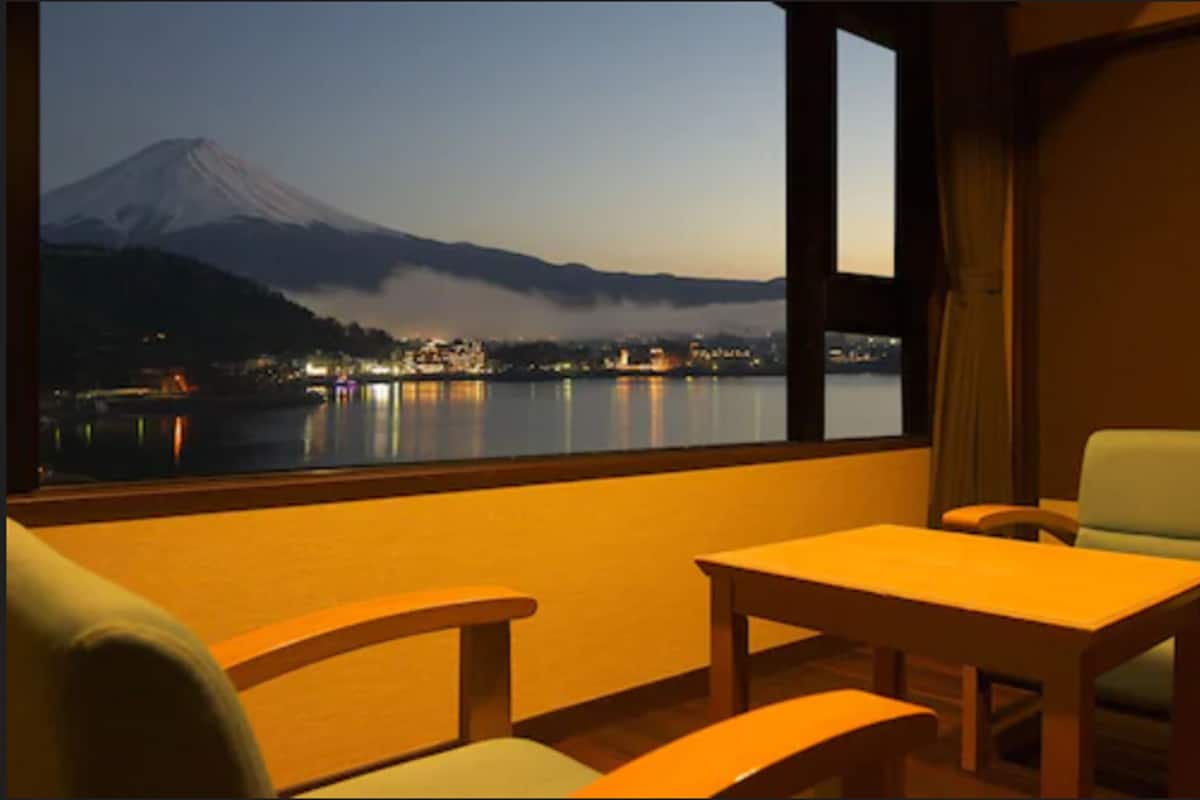 A room with a view of a mountain and a lake at dusk. There are two wooden tables and chairs.