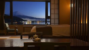 A hotel room with a bed, a dining table, and a view of a mountain and city lights. - Hotel Mifujien (Fujikawaguchiko)