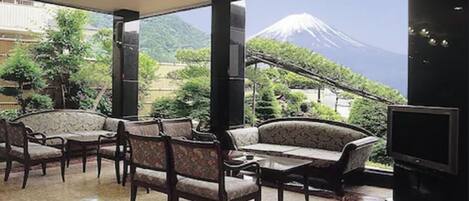 A hotel room with a large window offering a view of a mountain. Inside, there is a sofa, chairs, and a flat-screen TV.