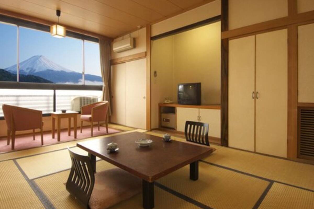 A traditional Japanese room with tatami flooring, a low table, chairs, a television, and a view of a mountain.