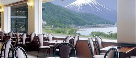 A restaurant with round tables and chairs, a view of a mountain, and a body of water.