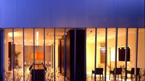 Un edificio moderno con ventanas amplias, un interior visible con asientos y un televisor de pantalla plana, y fachada de ladrillo.