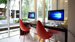 Una oficina moderna con dos escritorios, cada uno con un monitor de computadora, sillas rojas y ventanas amplias que ofrecen vistas a la calle y áreas verdes.