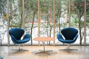 Una habitación moderna con dos sillas azules y una mesa redonda, ventanas amplias con vistas a árboles y edificios.