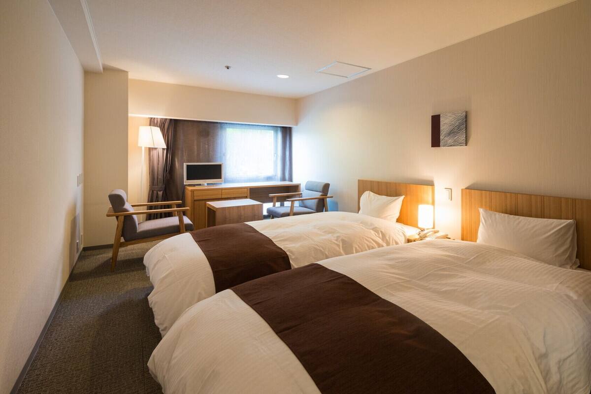 A hotel room with two beds, a desk, a chair, a television, and a window.