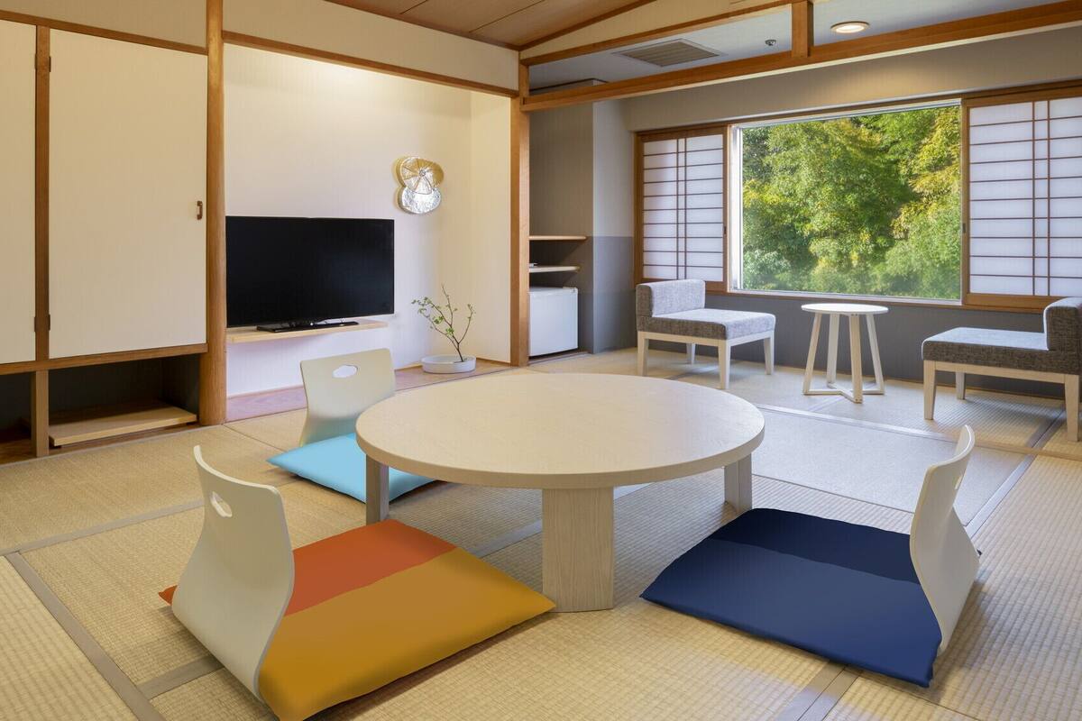 A traditional Japanese-style room with tatami flooring, a tatami mat table, low seating, a television, and a view of greenery through a shoji screen.