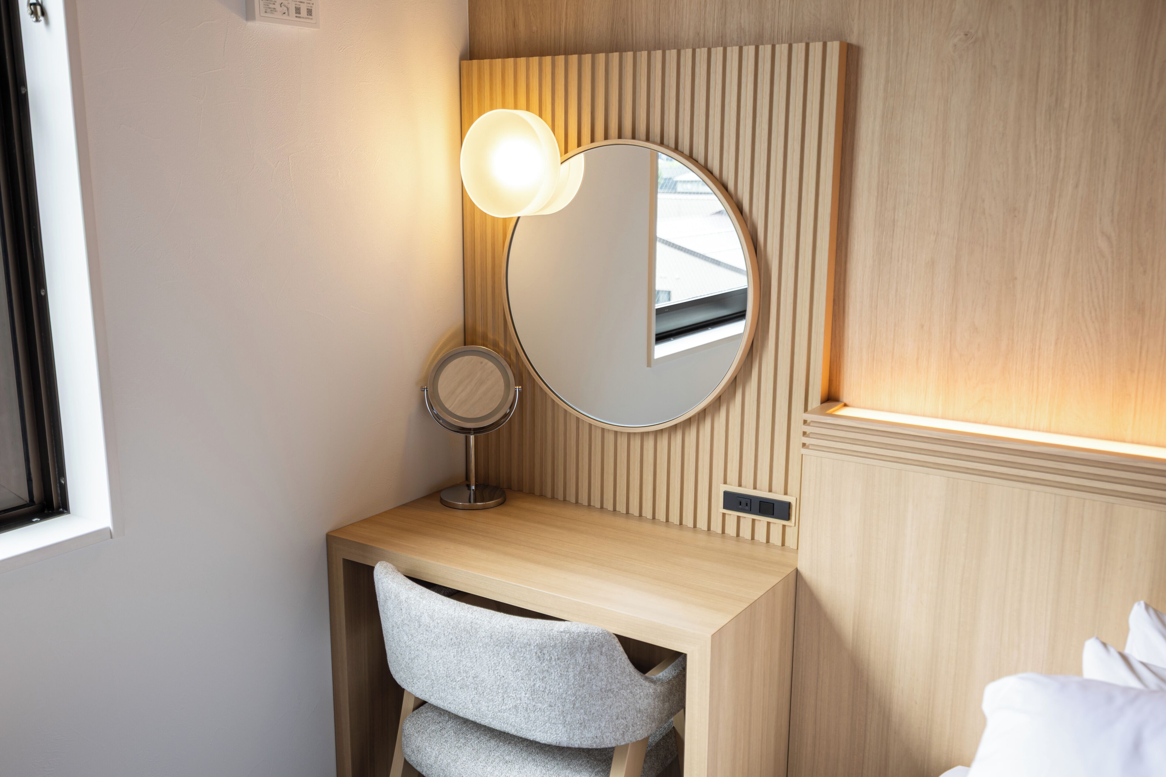 A wooden vanity with a round mirror and a light fixture, a chair, and a bed with white bedding.
