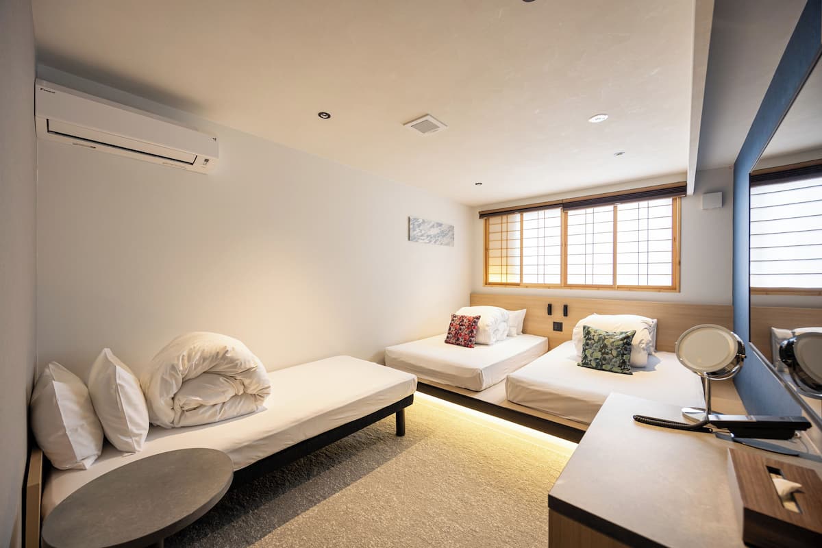 A hotel room with two single beds, a small table, a mirror, and a window with blinds.